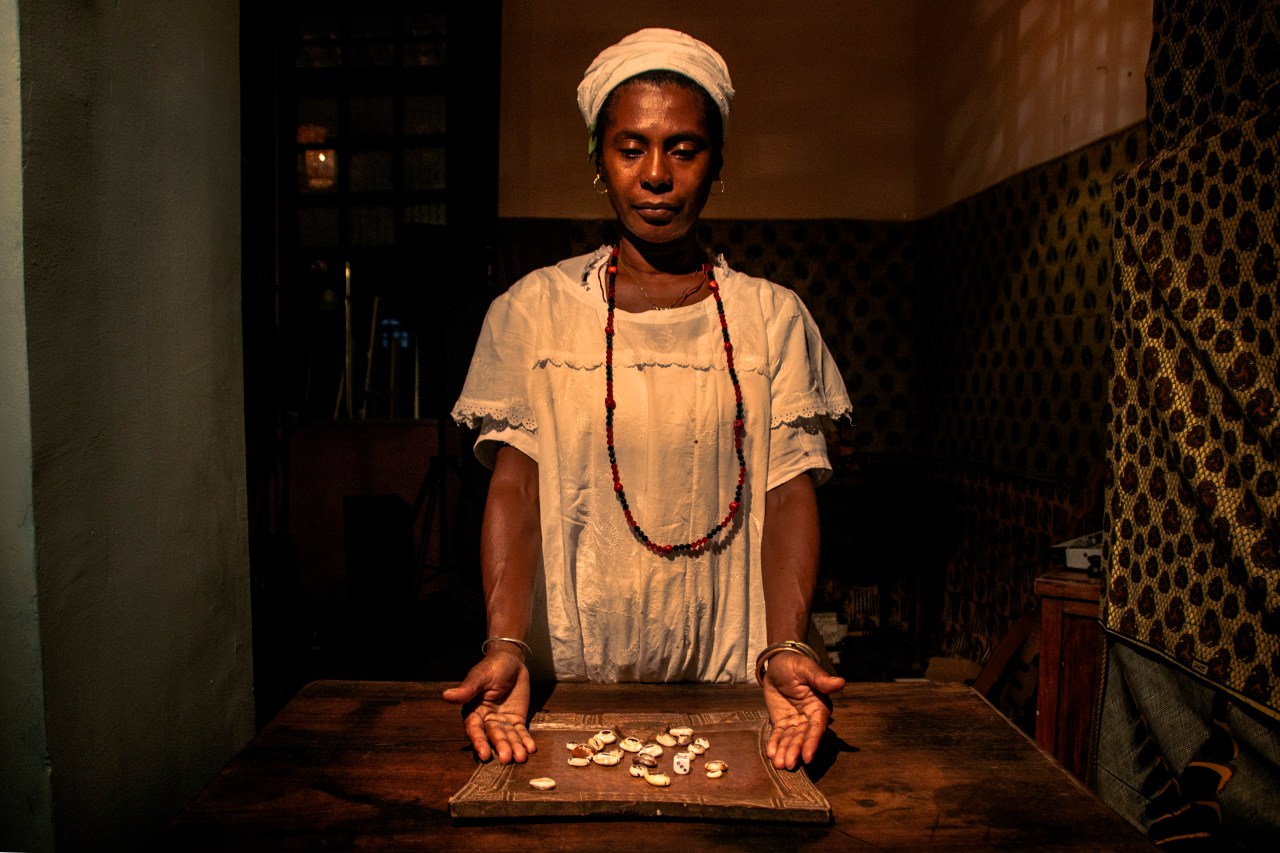 Mãe Marta stands above her divination instruments, the buzios or cowrie shells. As she tosses them on her table, the position in which they fall indicates a response from the Orishas, which she interprets for the inquirer.